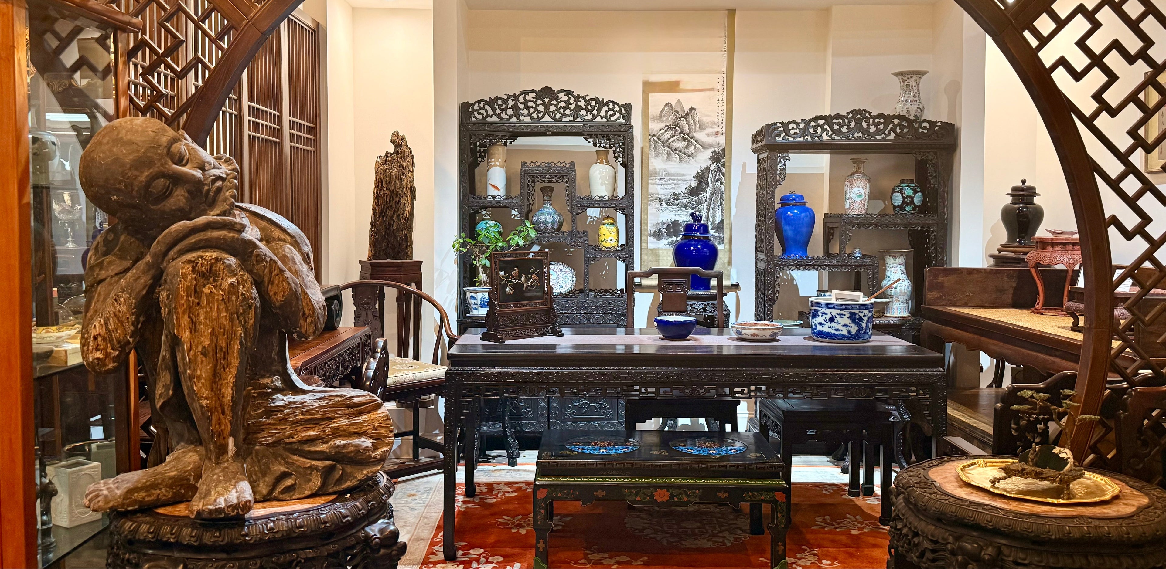 A traditional Chinese-style interior room with carved wooden furniture, an arched lattice screen, a large carved wooden statue in the foreground, and multiple display cabinets and mirrors.