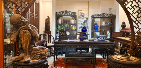 A traditional Chinese-style interior room with carved wooden furniture, an arched lattice screen, a large carved wooden statue in the foreground, and multiple display cabinets and mirrors.