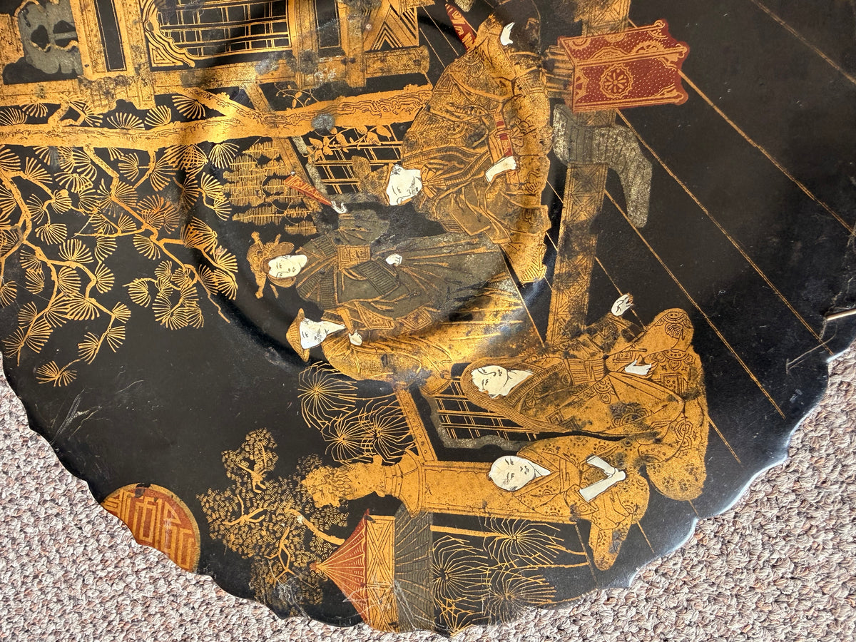 A close-up of a black lacquered decorative object featuring intricate gold and white Japanese-style painted figures and architectural details, photographed on a carpeted floor.