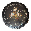 A round black lacquered tray with a scalloped edge, featuring a central painted floral bouquet and scattered iridescent shell-like flower petals on a white background.