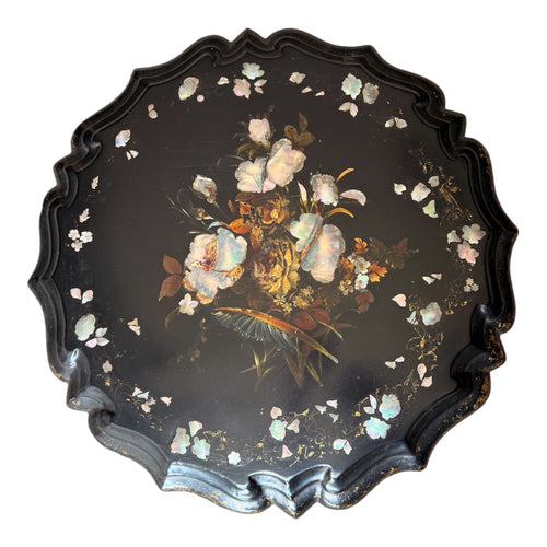 A round black lacquered tray with a scalloped edge, featuring a central painted floral bouquet and scattered iridescent shell-like flower petals on a white background.