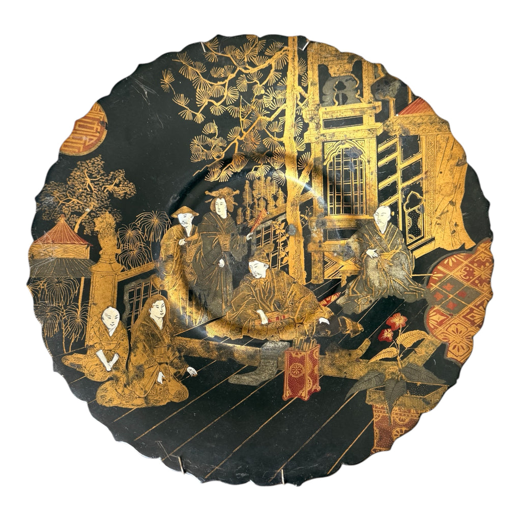 A round black lacquered plate with a scalloped edge, decorated with intricate gold and red hand-painted scenes of figures in traditional attire in an outdoor architectural setting.