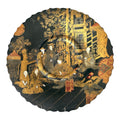 A round black lacquered plate with a scalloped edge, decorated with intricate gold and red hand-painted scenes of figures in traditional attire in an outdoor architectural setting.