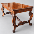 An ornate carved wooden table with a polished reddish-brown finish, featuring decorative legs and a rectangular top, photographed on a neutral light gray background.